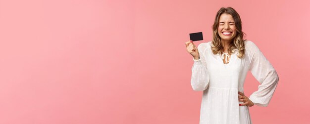 Retrato de una chica rubia emocionada, linda y tonta, vestida de blanco, con los ojos cerrados, riéndose y sonriendo feliz, recibió su primer pago, nuevo trabajo, con tarjeta de crédito, uso del servicio bancario, fondo rosa