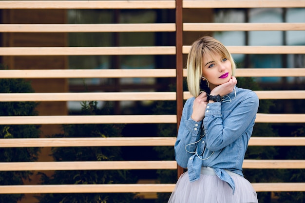 Un retrato de una chica rubia de ensueño con labios de color rosa brillante y maquillaje desnudo escuchando música en un teléfono inteligente con balks de madera a rayas detrás