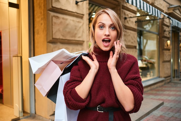 Retrato de una chica rubia con estilo sorprendida con bolsas de compras mirando asombrosamente en cámara en la calle de la ciudad
