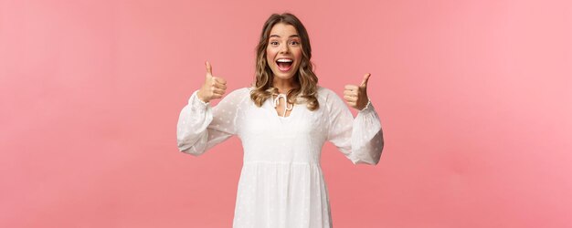 Retrato de una chica rubia femenina emocionada con vestido blanco muestra el pulgar hacia arriba en aprobación como si estuviera complacido asiente con la cabeza sonriendo fascinado deja una revisión positiva recomendar el producto fondo rosa