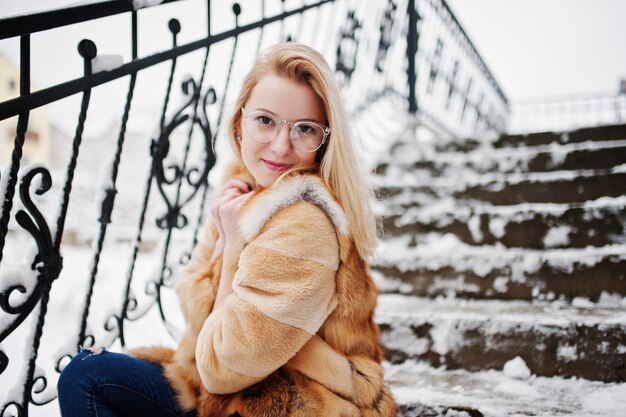 Retrato de una chica rubia con gafas, abrigo de piel rojo y bufanda en el día de invierno