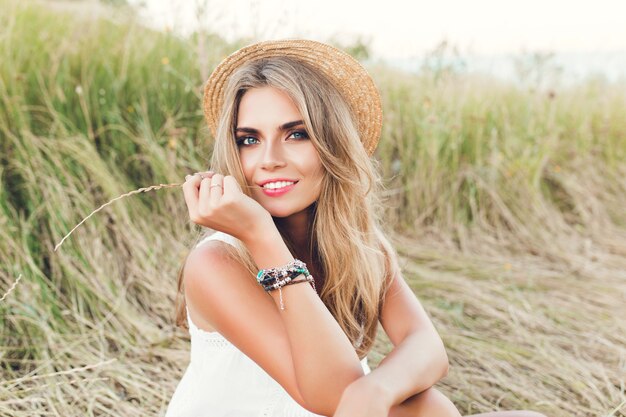 Retrato de chica rubia con pelo largo posando para la cámara sobre fondo de campo. Ella sostiene la hierba y mira a la cámara.