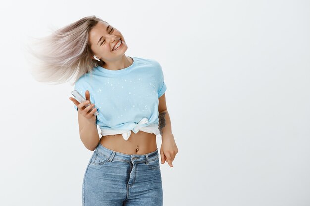 Retrato de chica rubia positiva posando en el estudio con su teléfono y auriculares