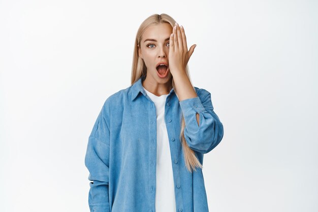 Retrato de una chica rubia que cubre un lado de la cara que parece impresionada con un ojo jadeando de asombro de pie contra el fondo blanco