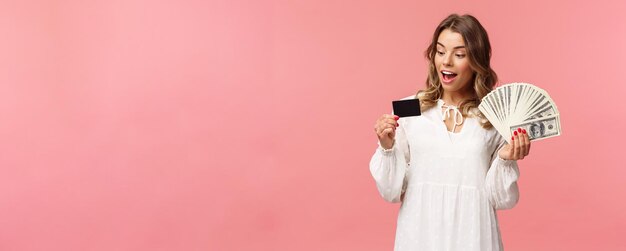 Retrato de una chica rubia rica y guapa con vestido blanco sosteniendo dólares y ganando con tarjeta de crédito