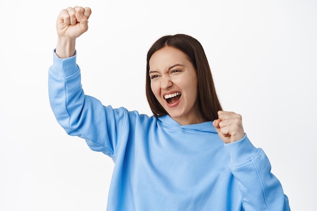 Retrato de una chica sonriente emocionada celebrando, gritando y cantando, haciendo puñetazos, mirando a un lado animando al equipo, animando y regocijándose, de pie contra el fondo blanco