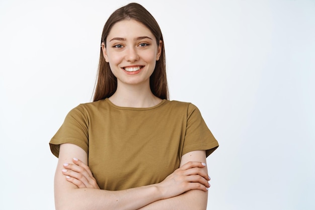 Retrato de una chica sonriente y segura de sí misma cruza los brazos sobre el pecho de pie en una pose profesional real con una camiseta informal de fondo blanco