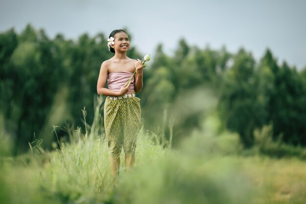 Retrato de chicas encantadoras en traje tradicional tailandés y poner flor blanca en su oreja, de pie y sosteniendo dos loto en la mano en el campo de arroz, sonríe con felicidad, copia espacio