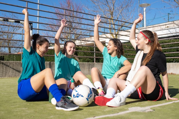 Retrato de chicas felices hablando en el campo de fútbol. Cuatro lindas chicas caucásicas con ropa deportiva sentadas en el suelo, levantando las manos para demostrar su fe en la victoria. Concepto de estilo de vida saludable y deporte de equipo