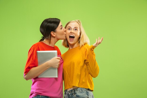 Retrato de chicas muy encantadoras en trajes casuales aislados en la pared verde. Novias o lesbianas que usan una tableta para divertirse o hacer pagos. Concepto de LGBT, emociones humanas, amor, relación.