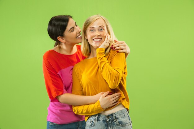 Retrato de chicas muy encantadoras en trajes casuales aislados sobre fondo verde de estudio. Dos modelos femeninas como novias o lesbianas. Concepto de LGBT, igualdad, emociones humanas, amor, relación.