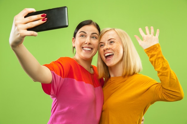 Retrato de chicas muy encantadoras en trajes casuales aislados sobre fondo verde de estudio. Novias o lesbianas haciendo selfie. Concepto de LGBT, igualdad, emociones humanas, amor, relación.