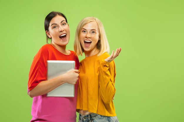 Retrato de chicas muy encantadoras en trajes casuales aislados sobre fondo verde de estudio. Novias o lesbianas que usan una tableta para divertirse o hacer pagos. Concepto de LGBT, emociones humanas, amor, relación.