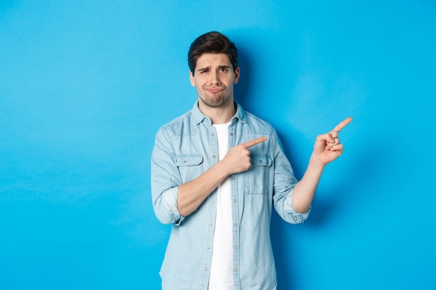 Retrato de chico adulto escéptico señalando con el dedo a la derecha y sonriendo, expresando decepción y duda, de pie contra el fondo azul.