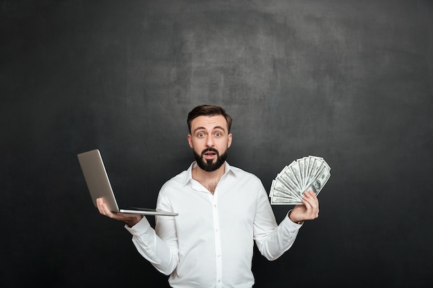 Retrato de chico adulto sorprendido en camisa blanca con abanico de billetes de dólar de dinero y cuaderno de plata en ambas manos sobre gris oscuro