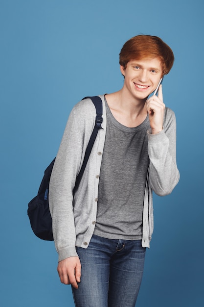 Retrato de chico alegre joven estudiante atractivo con el pelo de jengibre en traje casual con mochila, sonriendo alegremente, hablando con un amigo en el teléfono celular con expresión feliz.