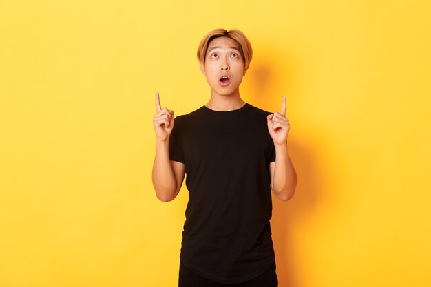Retrato de chico asiático curioso y asombrado con cabello rubio, vestido con camiseta negra, mirando y señalando con el dedo hacia arriba, pared amarilla asombrada