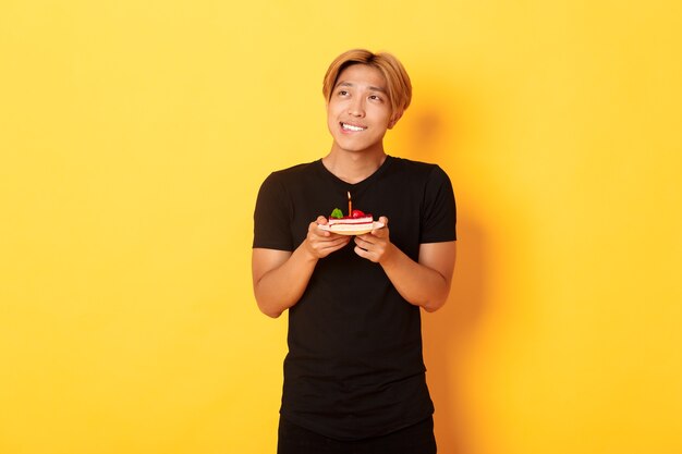 Retrato de un chico asiático de ensueño guapo mirando la esquina superior izquierda y pensando, pidiendo un deseo mientras celebra el cumpleaños y sostiene el b-day cake, pared amarilla