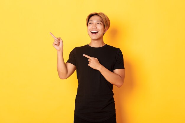 Retrato de chico asiático guapo complacido en camiseta negra, señalando con el dedo y mirando en la esquina superior izquierda con sonrisa satisfecha, pared amarilla