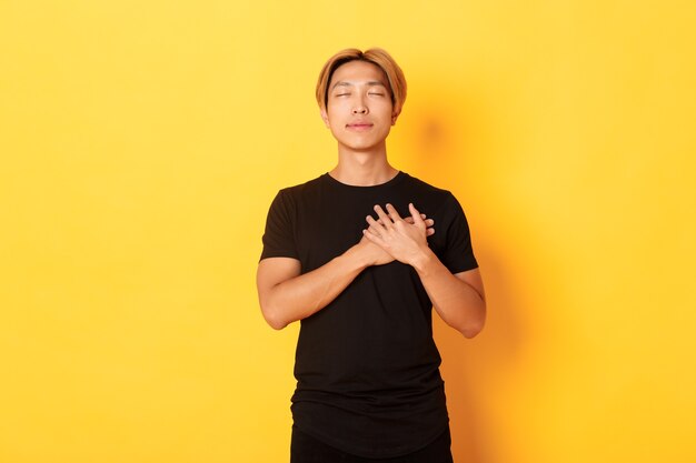 Retrato de chico asiático guapo tocado tomados de la mano en el corazón y sonriendo halagado, pared amarilla