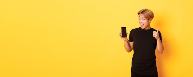 Foto gratuita retrato de un chico asiático satisfecho mirando la pantalla del teléfono inteligente y mostrando el pulgar hacia arriba en standi de aprobación
