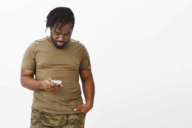 Retrato de chico atractivo en una camiseta marrón posando contra la pared blanca con su teléfono