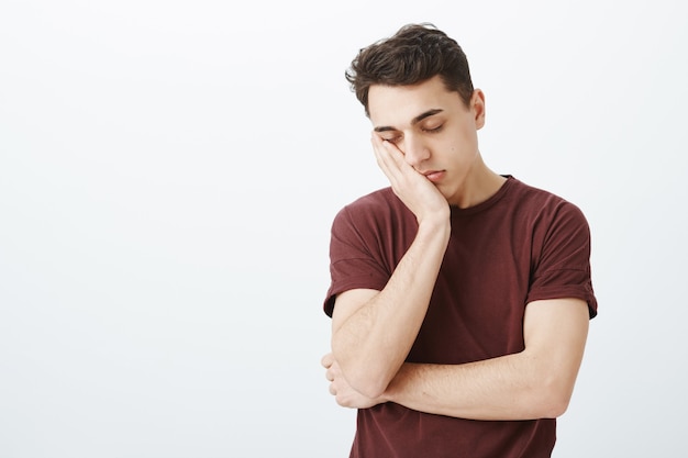 Retrato de un chico encantador y soñoliento con ropa casual, con la cara apoyada en la palma y cerrando los ojos, sintiéndose cansado y agotado, sin dormir