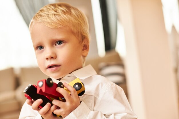 Retrato de chico europeo rubio guapo posando en el interior de la elegante sala de estar con camisa blanca disfrutando de juegos de interior jugando coloridos carros o coches. Creatividad, imaginación y fantasía