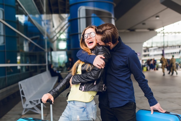 Retrato de chico guapo con barba en camisa negra besando a chica con pelo largo fuera en el aeropuerto. Lleva gafas, suéter amarillo y chaqueta con jeans. Ella luce feliz.