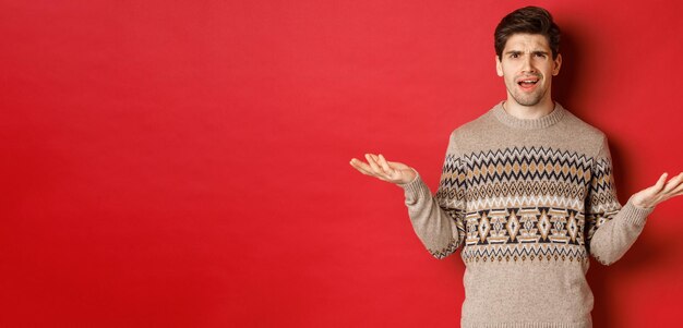 Retrato de un chico guapo confundido y decepcionado, quejándose de la Navidad, extendiendo las manos a los lados y frunciendo el ceño disgustado, parado en un suéter de Navidad sobre fondo rojo.