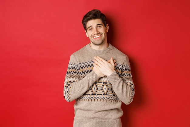 Retrato de un chico guapo conmovido y feliz, recibiendo un regalo de año nuevo, tomados de la mano en el corazón y sonriendo, diciendo gracias, de pie en un suéter de Navidad contra el fondo rojo.