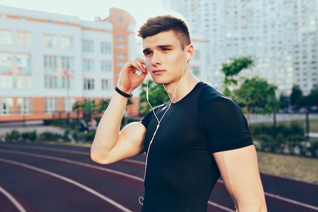 Retrato de chico guapo con un cuerpo musculoso por la mañana en el estadio. Viste camiseta negra, se pone los auriculares en la oreja y mira a un lado.