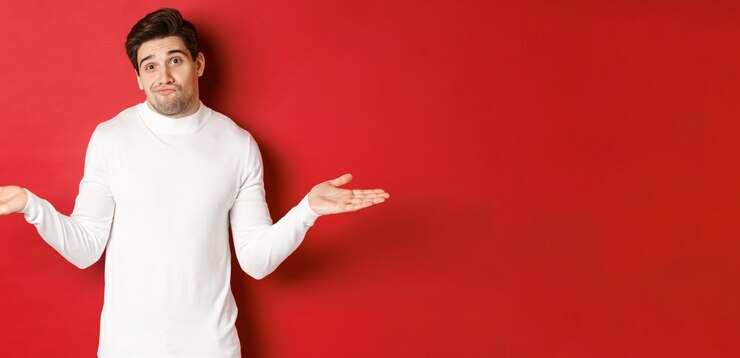 Foto gratuito retrato de un chico guapo despistado encogiéndose de hombros con las manos extendidas de lado sin darse cuenta de que no sabe nada de pie sobre un fondo rojo