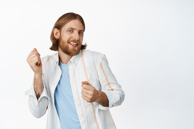 Retrato de chico guapo rubio barbudo, bailando despreocupado, sonriendo alegre, de pie en traje casual en blanco. Copia espacio