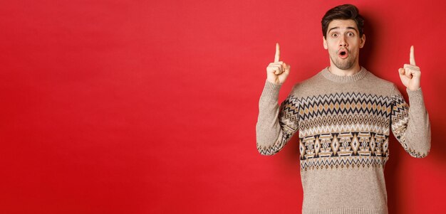 Retrato de un chico guapo sorprendido y asombrado que muestra una oferta promocional de navidad, usa un suéter de navidad y se para sobre un fondo rojo
