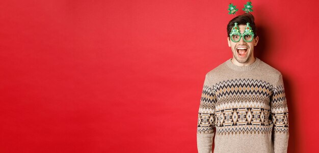 Retrato de un chico guapo sorprendido y emocionado con gafas de fiesta y suéter de invierno celebrando la Navidad y divirtiéndose de pie contra el fondo rojo