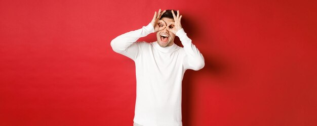 Retrato de chico guapo en suéter blanco haciendo máscara divertida con los dedos mirando feliz y sonriente s