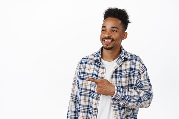 Retrato de chico negro feliz, joven barbudo de 20 años sonriendo satisfecho, señalando con el dedo hacia la izquierda y mirando el banner de venta, mostrando el logo a un lado en blanco. Copia espacio