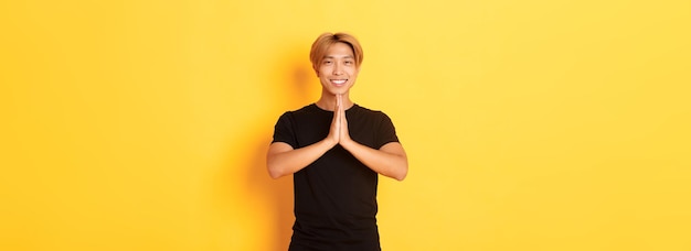 Foto gratuita retrato de un chico rubio asiático sonriente y agradecido cogidos de la mano juntos en oración de pie backgrou amarillo