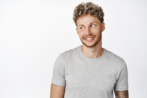 Retrato de un chico rubio atractivo sonriendo mirando a un lado el espacio de la copia usando una camiseta gris de pie contra el fondo blanco