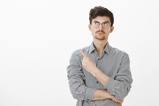 Retrato de un chico tranquilo, intrigado y vacilante con barba y bigote con gafas redondas, mirando y señalando la esquina superior izquierda con expresión curiosa, viendo algo interesante sobre una pared gris