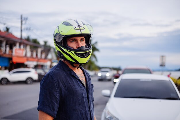 Retrato de ciclista masculino en casco amarillo al lado de una carretera muy transitada en Tailandia al atardecer