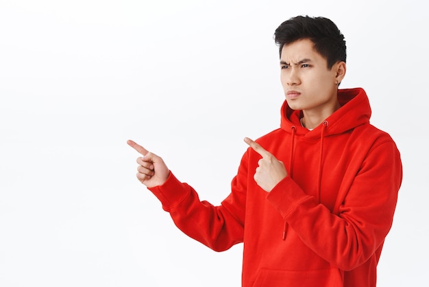 Foto gratuita retrato en la cintura de un apuesto hombre asiático que parece crítico o con odio, entrecerrando los ojos, frunciendo el ceño, señalando y mirando con ira y desprecio a la envidia de su enemigo de fondo blanco