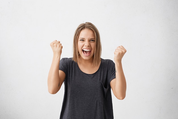 Retrato de cintura para arriba de la ganadora joven decidida y exitosa fuerte en camiseta casual levantando los brazos, apretando los puños, exclamando con alegría y emoción. Concepto de victoria, éxito y logro