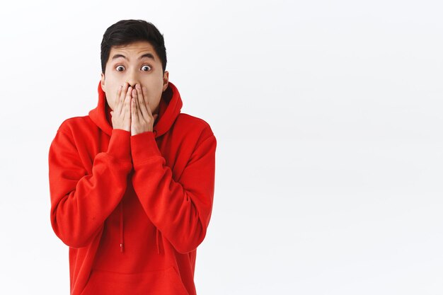 Retrato de cintura para arriba de un hombre asiático mudo conmocionado, jadeando, tapando la boca y mirando a la cámara, expresando temblar con los ojos, chismeando, escuchado emocionantes noticias increíbles