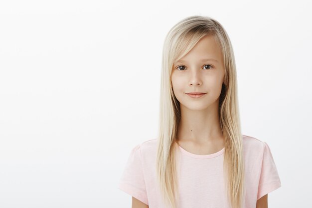 Retrato de cintura para arriba de una linda niña relajada con cabello rubio natural en camiseta rosa, de pie seguro y relajado sobre una pared gris, sonriendo levemente, de buen humor, esperando en la cola