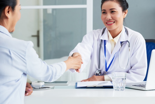 Retrato en la cintura del médico asiático estrechándole la mano con el paciente irreconocible sentado de espaldas a la cámara