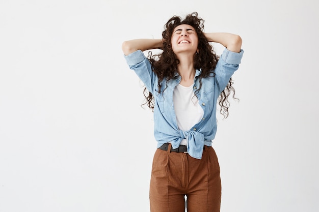 El retrato a la cintura de la modelo femenina de cabello oscuro emocional que usa una camisa vaquera aprieta sus dientes blancos y llenos de alegría, con los ojos cerrados y las manos sobre su cabeza. Expresión facial, emociones y sentimientos.