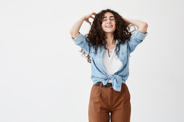 El retrato a la cintura de la modelo femenina emocional positiva, morena, viste camisa vaquera, sonríe ampliamente, juega con el cabello largo y ondulado, se alegra de la vida, muestra dientes blancos. Belleza, felicidad y juventud