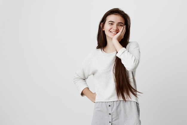 Retrato en la cintura de la modelo femenina morena sonriendo alegremente con dientes blancos, contento de recibir buenas noticias o presentes. La mujer emocionada expresa emotinos positivos, sostiene la mano en la mejilla con asombro.